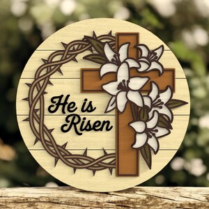 May include: Circular wooden Easter decoration with a brown cross, white lilies, and a crown of thorns. The words "He is Risen" are in black script. The background is a blurred green.