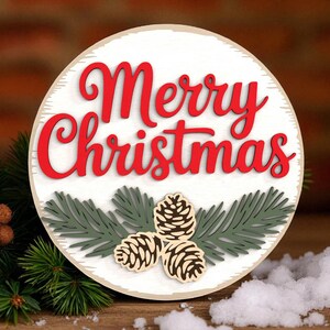 Pode incluir: Uma placa redonda de madeira branca com as palavras "Merry Christmas" em letras vermelhas. A placa apresenta agulhas de pinheiro verdes e três pinhas de madeira. Decoração festiva de férias.