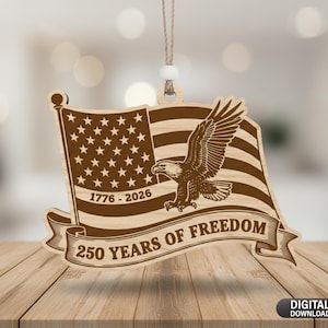 Op de afbeelding: Houten ornament met een ontwerp van de Amerikaanse vlag, een adelaar en de tekst "250 YEARS OF FREEDOM" en "1776-2026". Het ornament hangt aan een touwtje en wordt gepresenteerd op een houten oppervlak.