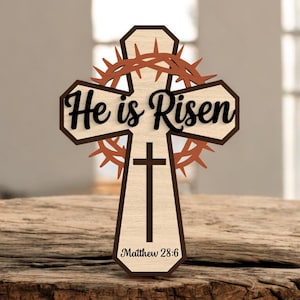 May include: Wooden cross with the words "He is Risen" in black script. A brown crown of thorns is above the cross, and the text "Matthew 28:6" is below. The cross is light brown with a dark brown border.