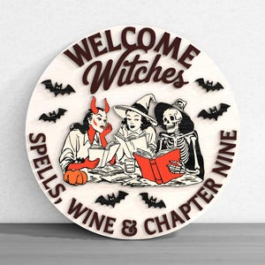May include: Round wooden sign with the words "WELCOME Witches" and "SPELLS, WINE & CHAPTER NINE". The sign features illustrations of a devil, a witch, and a skeleton reading books, with bats scattered around the edge. The sign is painted in shades of brown, red, and black.