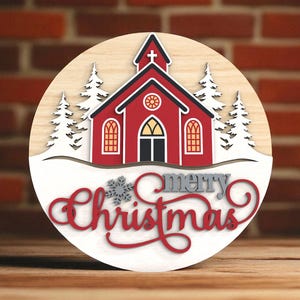 Puede incluir: Decoraci&oacute;n navide&ntilde;a redonda de madera con una iglesia roja, nieve blanca y &aacute;rboles. Las palabras "merry Christmas" est&aacute;n en escritura roja y gris. El fondo es una pared de ladrillos.