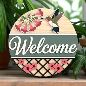 May include: A round wooden welcome sign with a hummingbird and flowers. The sign has a light wood top section, a green middle section with the word "Welcome" in white, and a black and wood lattice bottom with pink flowers.