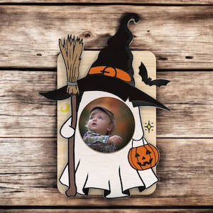 May include: Wooden Halloween-themed photo frame in the shape of a ghost wearing a witch hat. The frame has a circular opening for a photo, a broom, and a jack-o'-lantern bag. The witch hat is black with an orange band and a bat.