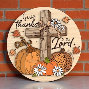 May include: A round wooden sign with a cross, pumpkins, and daisies. The sign reads "Give thanks to the Lord" with "Psalm 136:1" below. The pumpkins are orange and the daisies are white.