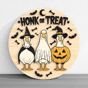 Puede incluir: Un letrero redondo de madera con tres gansos disfrazados para Halloween. Uno lleva un sombrero de bruja, otro una s&aacute;bana de fantasma y el tercero un disfraz de calabaza. El letrero dice "HONK OR TREAT" con murci&eacute;lagos, huesos y una luna.