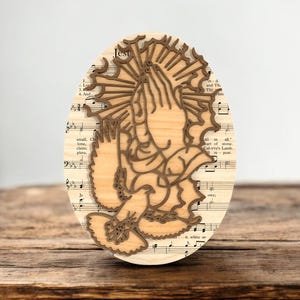 May include: Oval wooden art piece with a laser-cut design of praying hands and a halo. The background is sheet music. The art piece is made of light-coloured wood with a darker brown outline of the praying hands and halo.