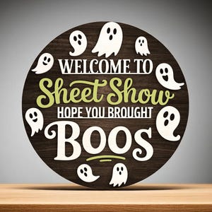 Puede incluir: Un letrero redondo de madera con las palabras "WELCOME TO Sheet Show HOPE YOU BROUGHT BOOS" en letras blancas y verdes. Siluetas de fantasmas blancos rodean el texto sobre un fondo de vetas de madera marr&oacute;n oscuro. El letrero est&aacute; sobre una superficie de madera.