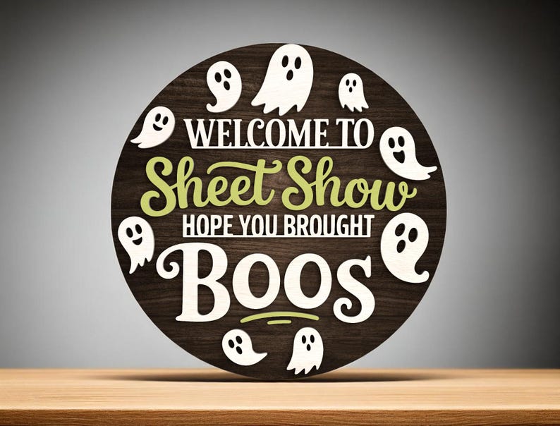 Puede incluir: Un letrero redondo de madera con las palabras "WELCOME TO Sheet Show HOPE YOU BROUGHT BOOS" en letras blancas y verdes. Siluetas de fantasmas blancos rodean el texto sobre un fondo de vetas de madera marr&oacute;n oscuro. El letrero est&aacute; sobre una superficie de madera.
