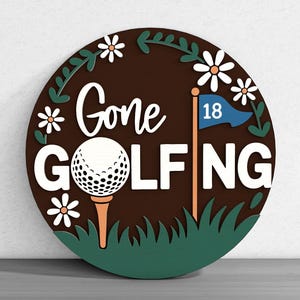 May include: A round wooden sign with the words "Gone Golfing" in white, a golf ball on a tee, and a flag with the number 18. The sign is brown with green and white accents, including daisies and grass.