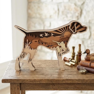 May include: A multi-layered wooden cutout in the shape of a hunting dog, featuring a laser-cut scene of a hunter with a rifle, a smaller dog, flying ducks, and a sunburst over a landscape. The natural wood tones create depth and contrast.