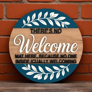 Puede incluir: Un letrero redondo de madera con el texto "THERE'S NO Welcome MAT HERE, BECAUSE NO ONE INSIDE ICUALLY WELCOMING". Cuenta con un borde azul con diseños de hojas blancas, sobre una pared de ladrillos.