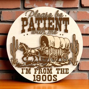 May include: Round wooden sign with a Western theme. The sign features a covered wagon pulled by horses, cacti, and the text "Please be PATIENT with me. I'M FROM THE 1900S" in brown lettering. The sign is displayed against a brick wall.