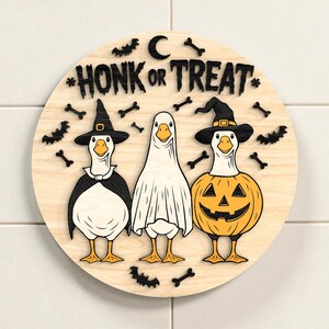 Puede incluir: Decoraci&oacute;n redonda de Halloween de madera con tres gansos disfrazados. Un ganso es una bruja, otro un fantasma y otro una calabaza. Las palabras "HONK OR TREAT" est&aacute;n en la parte superior, con murci&eacute;lagos y huesos.