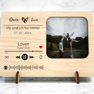Op de afbeelding: Houten fotolijst met een foto van een stel dat elkaars hand vasthoudt. De lijst heeft de namen "Davia" en "Anna" met een hartsymbool, de tekst "Du und ich fur immer", de datum "17-07-2014" en het nummer "Lover" van Taylor Swift.
