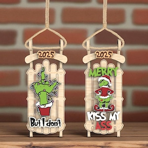 Puede incluir: Dos adornos navideños de madera con forma de trineo. Uno presenta una mano verde con luces navideñas y el texto "But I don't". El otro muestra un personaje verde con gorro de Papá Noel y el texto "Merry Kiss My Ass". Ambos tienen "2025" en la parte superior.
