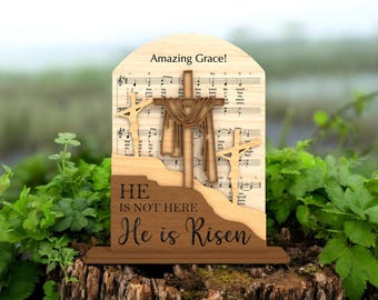 May include: Wooden religious decoration featuring three crosses, with the central cross draped with fabric. The background includes sheet music and the words "Amazing Grace!" The base reads "He is not here, He is Risen."