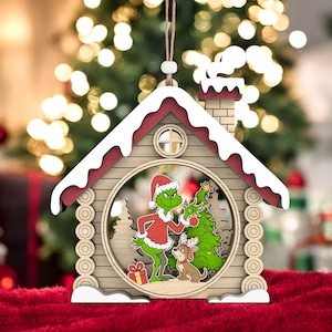 May include: Wooden Christmas house ornament with a green character in a Santa hat, a Christmas tree, and a dog. The house has a red roof with white snow and a chimney. The ornament hangs from a string. The background is blurred.