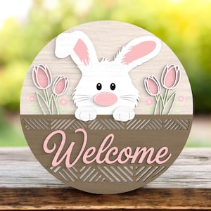 May include: A round wooden Easter-themed sign featuring a white bunny with pink ears and nose, peeking over a brown basket with the word "Welcome" in pink script. Pink tulips and small flowers are also present.