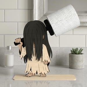 Puede incluir: Un portarrollos de papel higiénico de madera con una figura estilizada de pelo largo y negro y un vestido andrajoso. La figura sostiene una barra negra que sostiene un rollo de papel higiénico blanco. El soporte está sobre una base de madera.