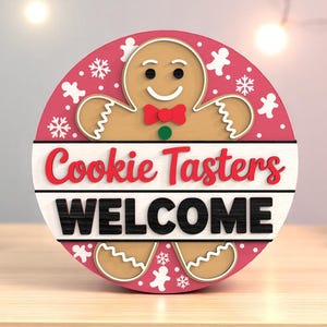 May include: A round, wooden sign with a gingerbread man design. The sign has a pink background with white snowflakes and gingerbread man cutouts. The words "Cookie Tasters WELCOME" are displayed in red and black.
