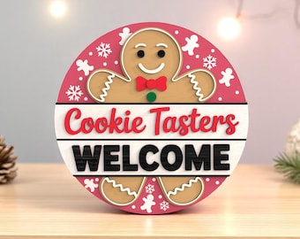 May include: A round, wooden sign with a gingerbread man design. The sign has a pink background with white snowflakes and gingerbread man cutouts. The words "Cookie Tasters WELCOME" are displayed in red and black.