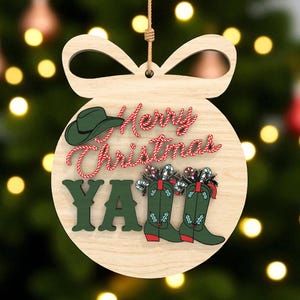 Puede incluir: Adorno navideño de madera con una parte superior en forma de lazo. El adorno presenta el texto "Merry Christmas Y'all" con un sombrero de vaquero y botas de vaquero. El texto es en rojo y verde, con bastones de caramelo en las botas.