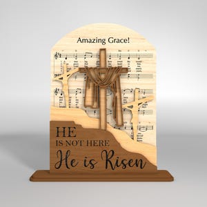 May include: Wooden Easter decoration featuring three crosses, with the central cross draped with fabric. The background includes sheet music and the words "Amazing Grace!" The base reads "He is not here, He is Risen."