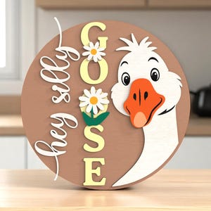 May include: A round wooden sign with a cartoon goose design. The sign has the words "hey silly goose" in white script and the word "GOOSE" in yellow block letters. Daisies are placed around the letters. The goose has a white head, orange beak, and black eyes.
