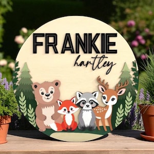 Peut inclure: Panneau rond en bois avec le nom "FRANKIE" en lettres noires au-dessus du nom "hartley" en cursive. Le panneau présente une scène de forêt avec un ours, un renard, un raton laveur et un cerf. Des arbres verts et du feuillage entourent les animaux.