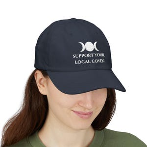 May include: Navy blue baseball cap with white embroidered design of three crescent moons and the text "SUPPORT YOUR LOCAL COVEN". The cap has a curved brim and a classic six-panel design. A stylish accessory for those interested in witchcraft and Wicca.