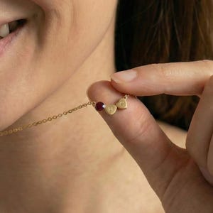 May include: A delicate gold necklace featuring a small, round, red gemstone and two heart-shaped charms with the letters "H" and "D". The necklace is held by a person's fingers.