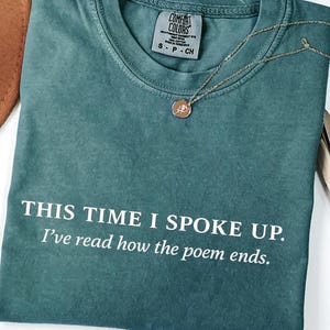 May include: A teal Comfort Colors t-shirt with the text "THIS TIME I SPOKE UP. I've read how the poem ends." A gold necklace with a pendant rests on the shirt. A brown hat is visible in the upper left corner.