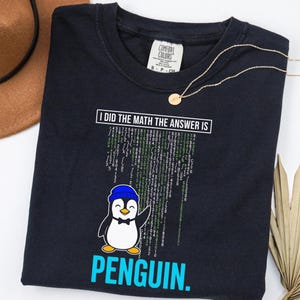 May include: Dark navy blue t-shirt with the text "I DID THE MATH THE ANSWER IS PENGUIN." A cartoon penguin wearing a blue hat and bow tie is featured below the text. The shirt also has a Comfort Colors tag.