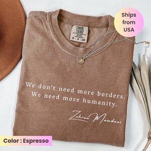 May include: Espresso-colored t-shirt with the text "We don't need more borders. We need more humanity." and the signature "Zohran Mamdani." A silver necklace with a pendant is on the shirt. The shirt is a Comfort Colors brand.