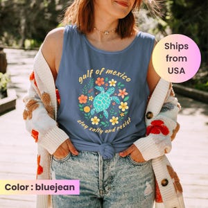 May include: Bluejean tank top with a turquoise sea turtle graphic surrounded by flowers and the text "gulf of mexico, stay salty and resist". The tank top is knotted at the waist and worn with light wash jeans and a cream-colored cardigan.