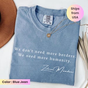 May include: A blue jean colored t-shirt with the text "We don't need more borders. We need more humanity." and the signature "Zohran Mamdani". The shirt has a crew neck and is paired with a gold necklace.