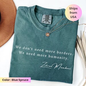 May include: Blue spruce Comfort Colors t-shirt with the text "We don't need more borders. We need more humanity." and the signature "Zohran Mamdani." A gold necklace with a pendant is on the shirt.