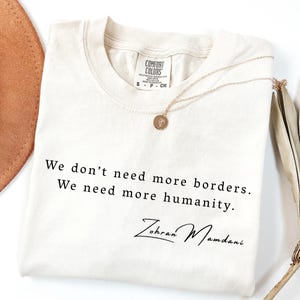 May include: Cream-colored t-shirt with the text "We don't need more borders. We need more humanity." and the signature "Zohran Mamdani" in black. A gold necklace with a small pendant rests on the shirt.
