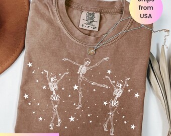 May include: Espresso-colored t-shirt featuring a white graphic of three dancing skeletons and scattered stars. The shirt has a crew neck and short sleeves. A silver necklace with a pendant is draped on the shirt.