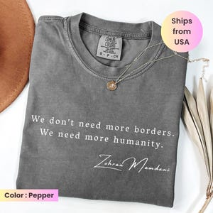 May include: A pepper-colored t-shirt with the text "We don't need more borders. We need more humanity. Zohran Mamdani" in white. A gold-colored necklace with a pendant rests on the shirt. The shirt is folded.
