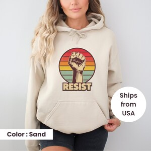 May include: A sand-colored hooded sweatshirt with a vintage-style graphic of a raised fist in front of a sunset. The graphic also includes the word "RESIST" in bold, black letters.
