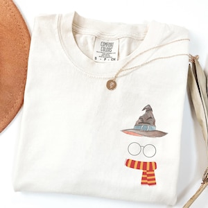 May include: An off-white short-sleeve t-shirt featuring a small graphic on the chest consisting of a brown pointed hat, round black-rimmed glasses, and a red and yellow striped scarf. The shirt has a Comfort Colors brand label at the neck.