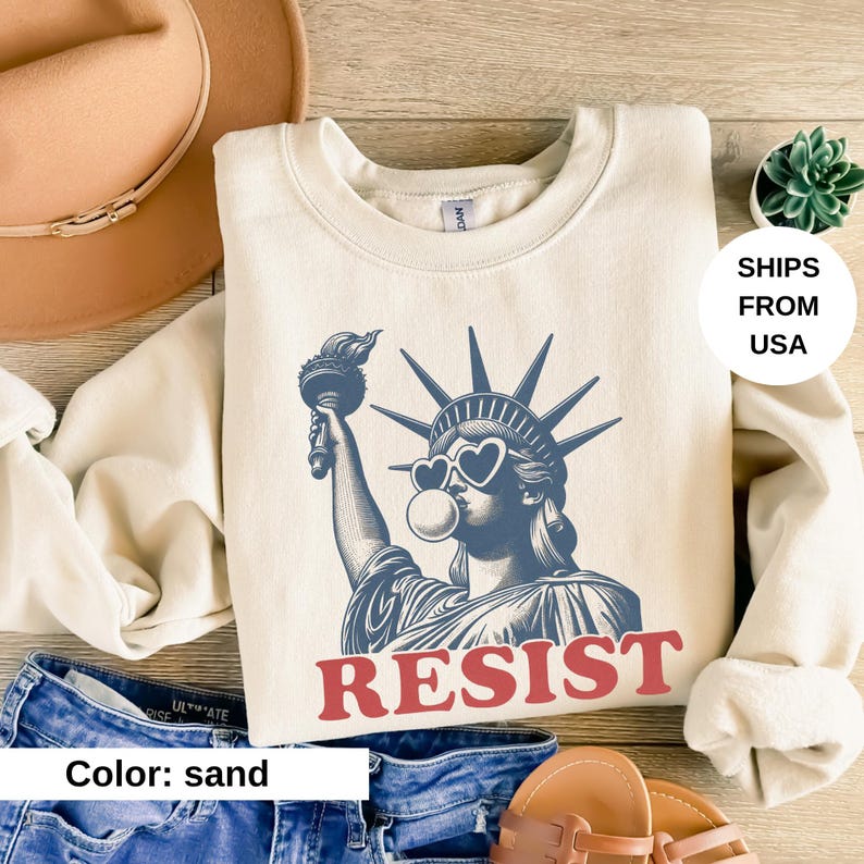 May include: Sand-coloured sweatshirt with a blue graphic of the Statue of Liberty wearing heart-shaped sunglasses and blowing a bubble. The word "RESIST" is printed in red below the graphic. A casual style sweatshirt.