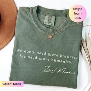 May include: Moss green Comfort Colors t-shirt with the text "We don't need more borders. We need more humanity." and the signature "Zohran Mamdani" in white. A gold necklace with a pendant is on the shirt.