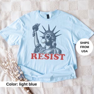 May include: Pale blue t-shirt with a graphic of the Statue of Liberty wearing heart-shaped sunglasses and blowing a bubble. The word "RESIST" is printed in red block letters below the image.