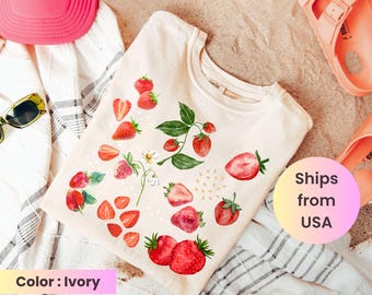 May include: Ivory-colored t-shirt with a watercolor strawberry design. The shirt features various illustrations of strawberries, including whole and sliced fruits, along with green leaves and small white flowers. The shirt is displayed on a beach towel with a pink hat and sandals.