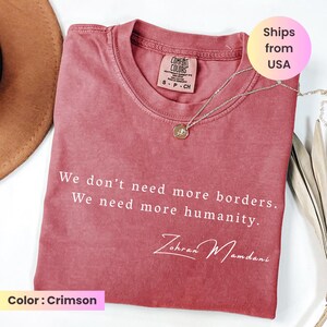 May include: Crimson-colored t-shirt with the text "We don't need more borders. We need more humanity." and the name "Zohran Mamdani" in white script. A silver necklace with a pendant is on the shirt.