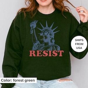 May include: Forest green sweatshirt with a blue graphic of the Statue of Liberty wearing heart-shaped sunglasses and blowing a bubble. The word "RESIST" is printed in red below the graphic.