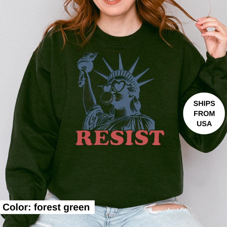 May include: Forest green sweatshirt with a blue graphic of the Statue of Liberty wearing heart-shaped sunglasses and blowing a bubble. The word "RESIST" is printed in red below the graphic.